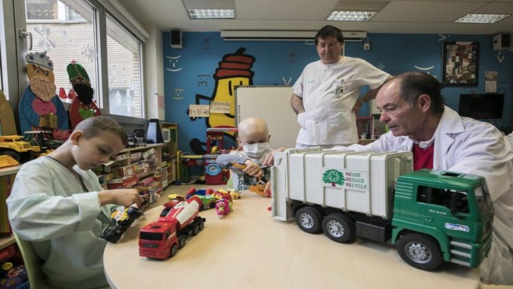 Un niño enfermo en el Aula Hospitalaria del Hospital Miguel Servet jugando con un camión de reciclaje junto a dos de los profesores del aula.