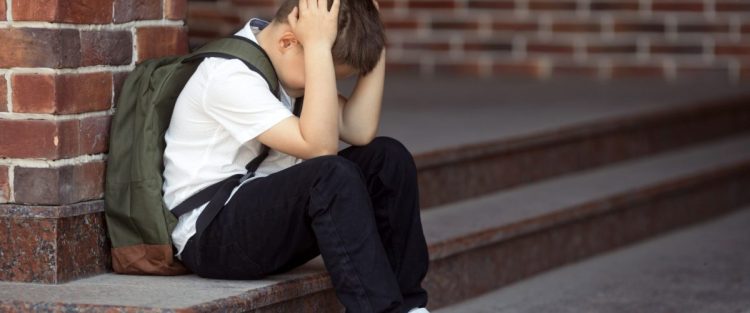 Niño víctima de acoso escolar. Está sentado en unas escaleras y tapándose la cabeza con las manos, cabizbajo.